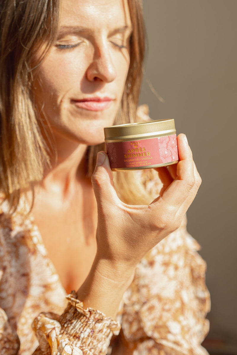 A person holds a small, round Apres Shimmer Holiday Tin Candle, evoking winter luxury with its scent of balsam fir and vanilla.