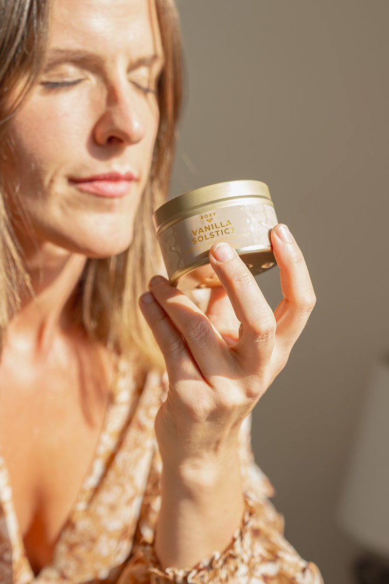 A person with eyes closed holds a small ROXY Vanilla Solstice Holiday Tin Candle up to their face.