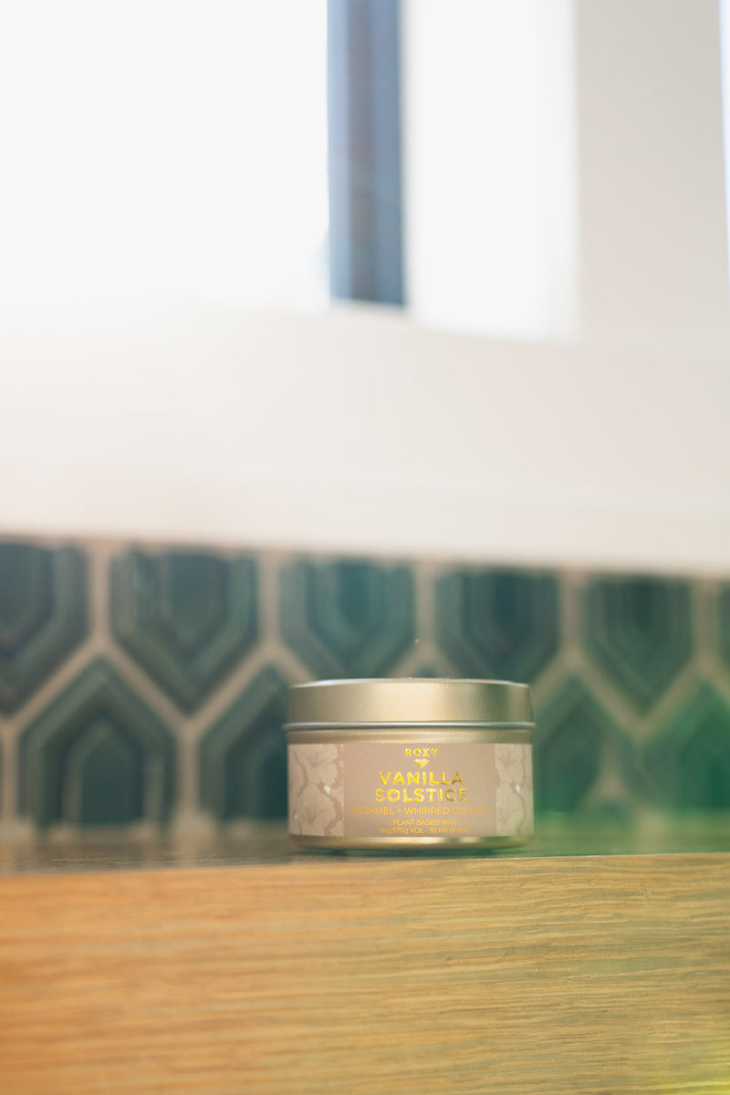 A cylindrical ROXY Vanilla Solstice Holiday Tin Candle with a floral pattern and gold text label sitting on a wooden surface.