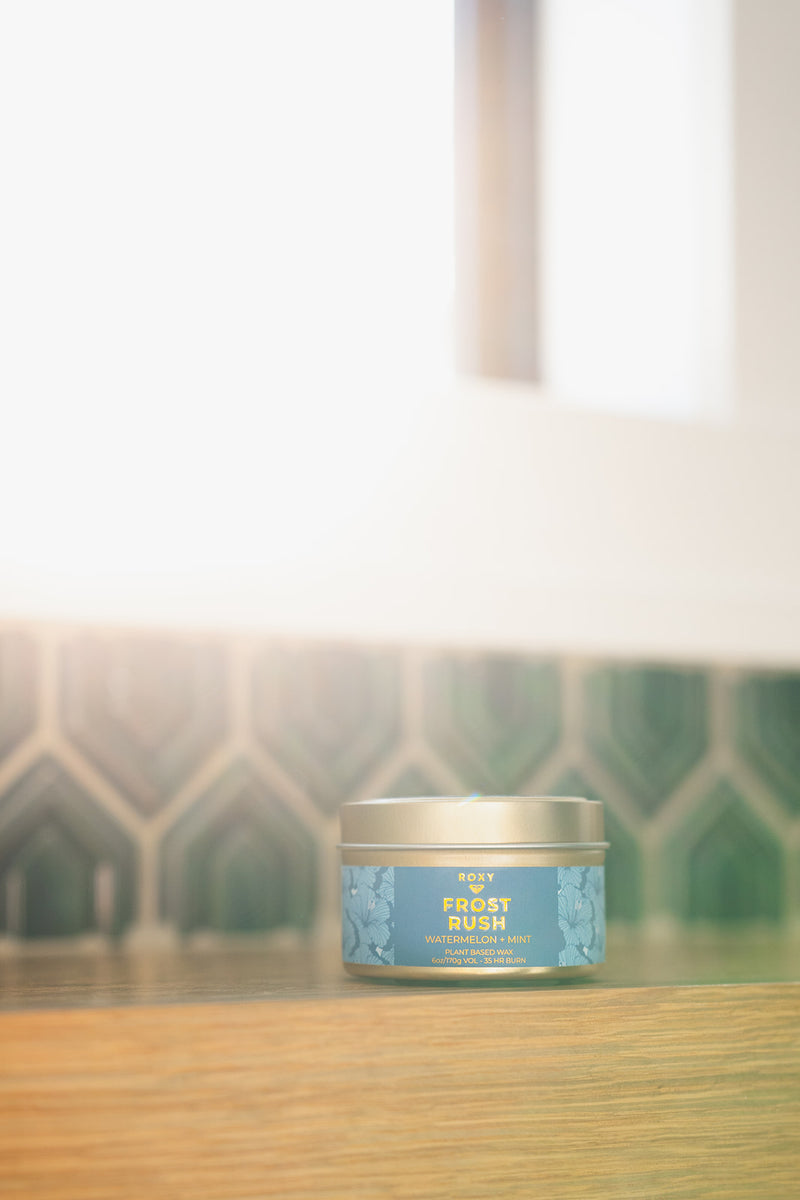 A small ROXY Frost Rush Holiday Tin Candle with crisp mint notes sits on a wooden shelf in front of a green tiled wall.