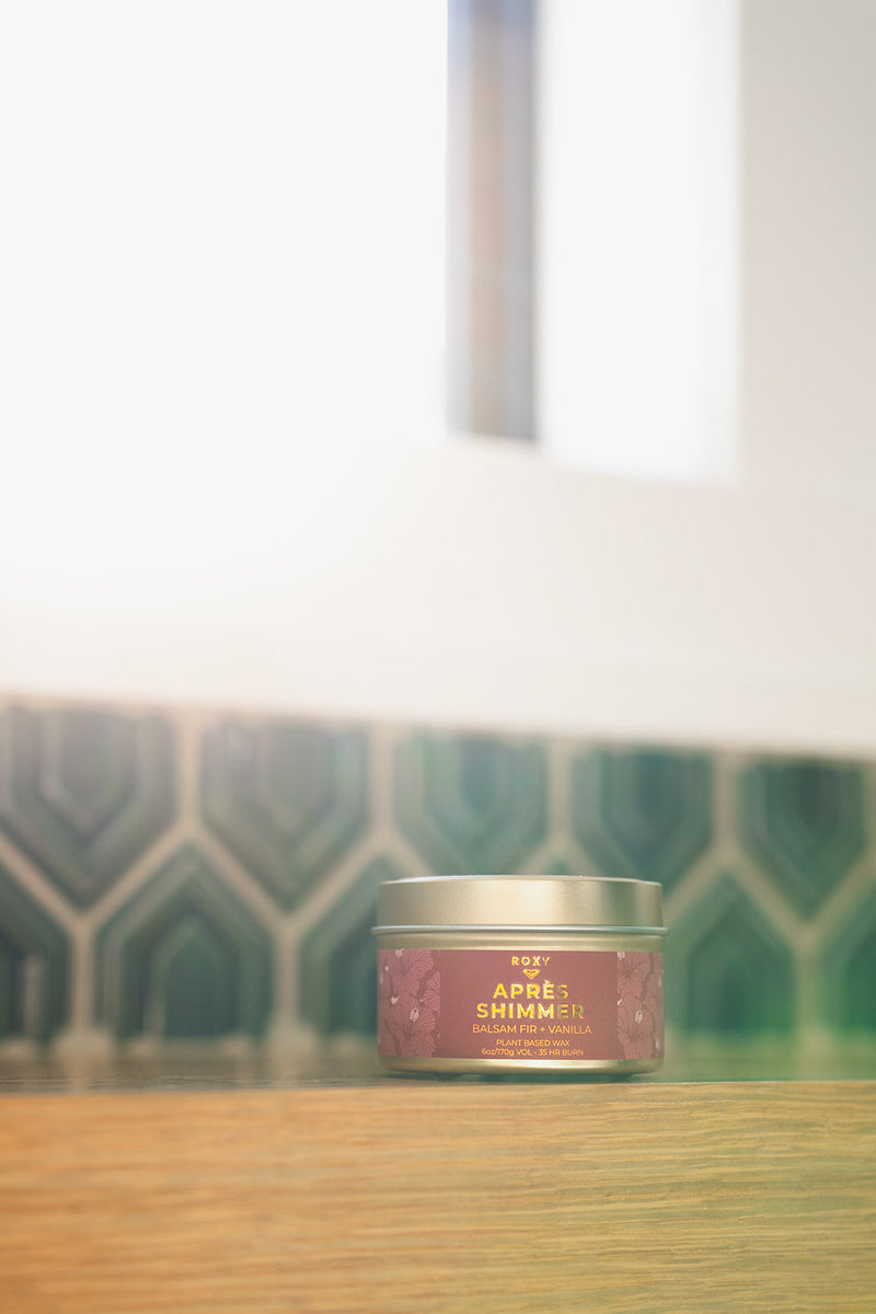 A round metal Apres Shimmer Holiday Tin Candle with a purple label featuring gold text reading Après Shimmer, Balsam Fir + Vanilla, evoking the essence of winter luxury.