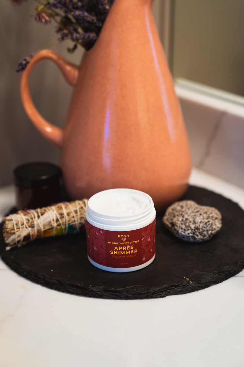 An open jar of ROXY Apres Shimmer Holiday Luxury Spa Set whipped body butter sits on a circular slate tray next to a smudge stick, a smooth stone, and a terracotta pitcher.