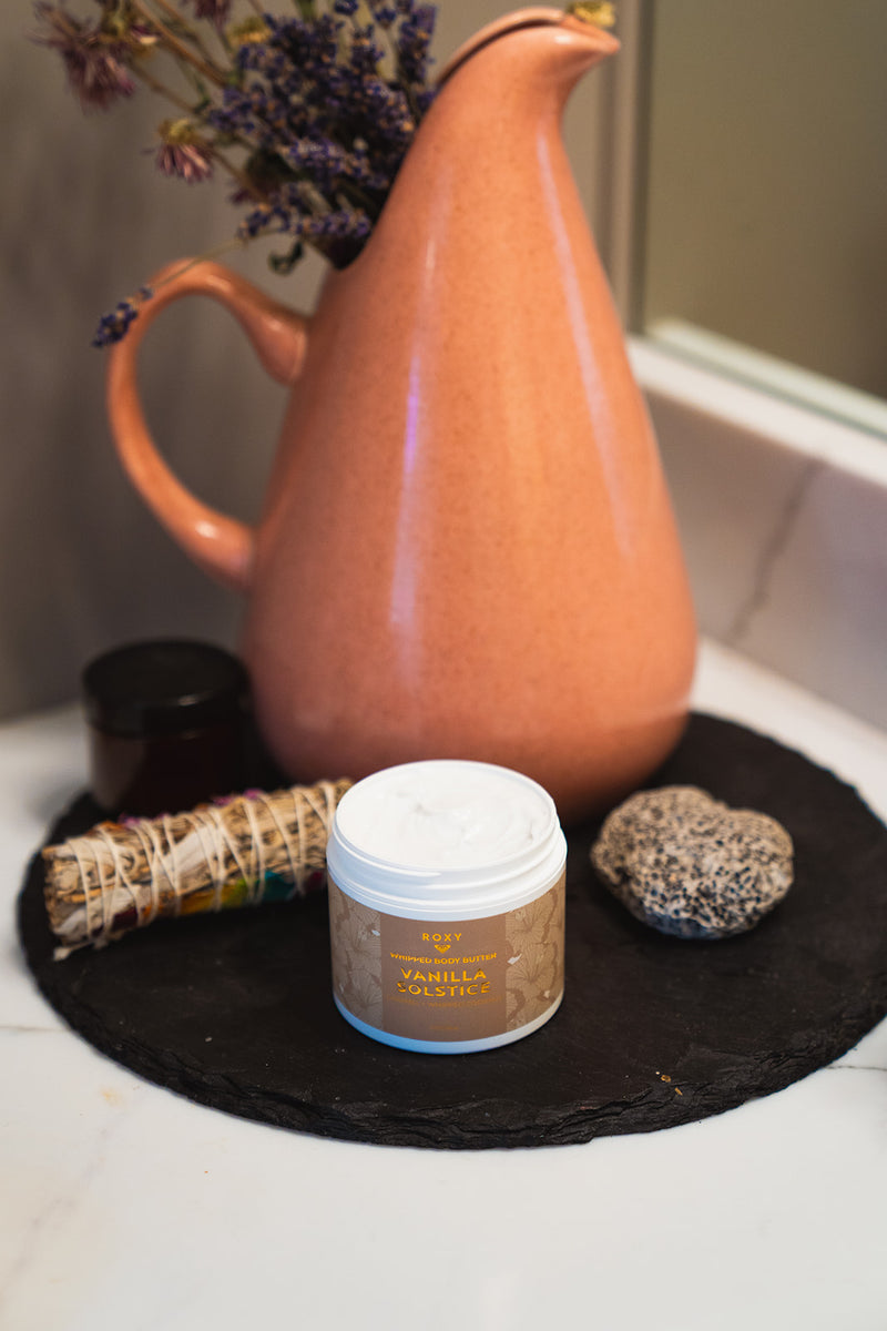 An open jar of Vanilla Solstice Holiday Luxury Spa Set body butter sits on a black circular stone tray next to a ceramic pitcher, a small jar, a bundle of dried herbs, and a smooth stone.