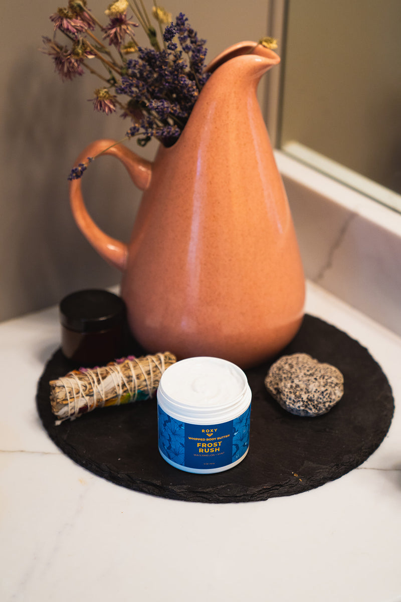 An open jar of Frost Rush Holiday Luxury Spa Set whipped body butter sits on a black circular tray beside a small dark jar, a sage bundle, a stone, and a pink pitcher filled with dried flowers.