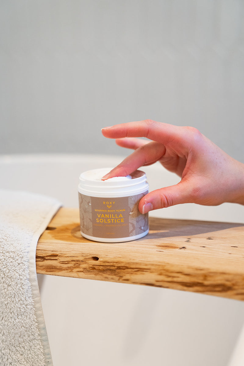 A hand reaches into an open jar of Vanilla Solstice Holiday Luxury Spa Set whipped body scrub resting on a wooden bath tray.
