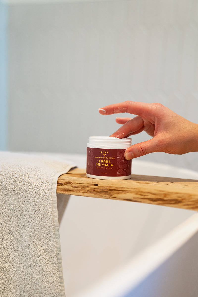 A hand reaches for a jar of Apres Shimmer Holiday Luxury Spa Set whipped body scrub, part of a calming body-care ritual, resting on a wooden bath tray.