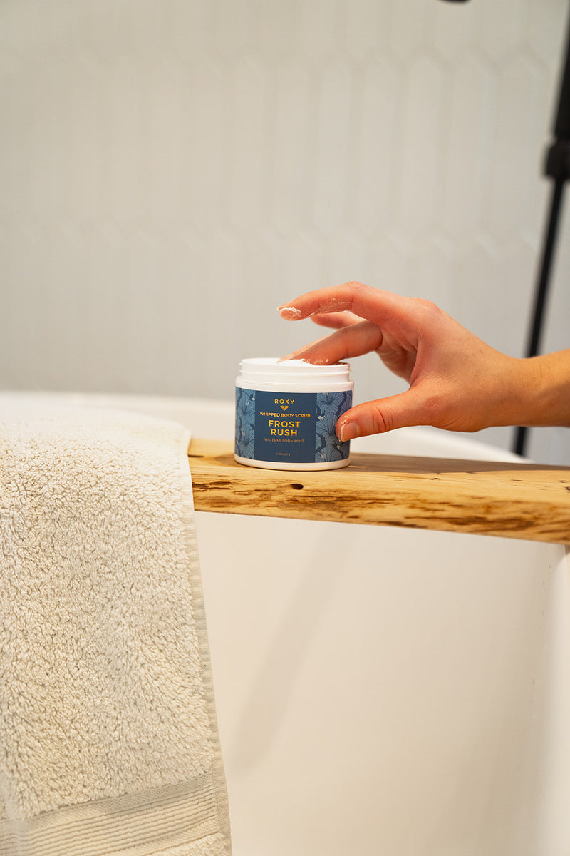 A hand reaches for a jar of whipped body scrub from a ROXY Frost Rush Holiday Luxury Spa Set resting on a wooden ledge above a bathtub.