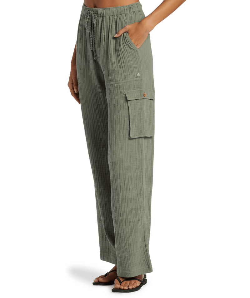 A person wears the Roxy Precious Cargo Slub Pant—loose-fitting, sage green with a drawstring waist and side pocket—paired with black sandals, standing against a plain white background.