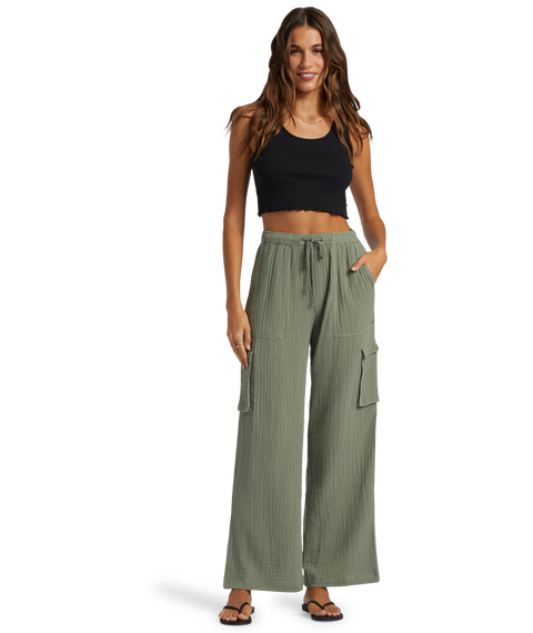 Woman wearing a black crop top, Roxy Precious Cargo Slub Pant in Agave Green Solid, and black sandals stands smiling with one hand in her pocket against a white background.