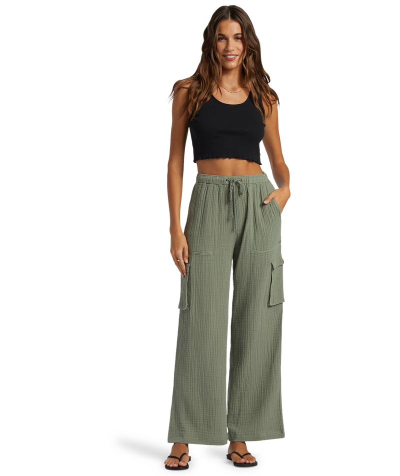 Woman wearing a black crop top, Roxy Precious Cargo Slub Pant in Agave Green Solid, and black sandals stands smiling with one hand in her pocket against a white background.