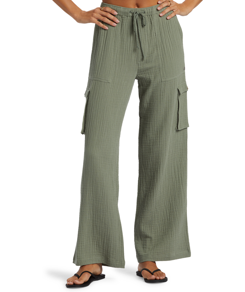 A person wears the Roxy Precious Cargo Slub Pant in sage green with a drawstring waist and side pockets, styled with black flip-flops. The photo shows the pants from waist to feet against a plain background.