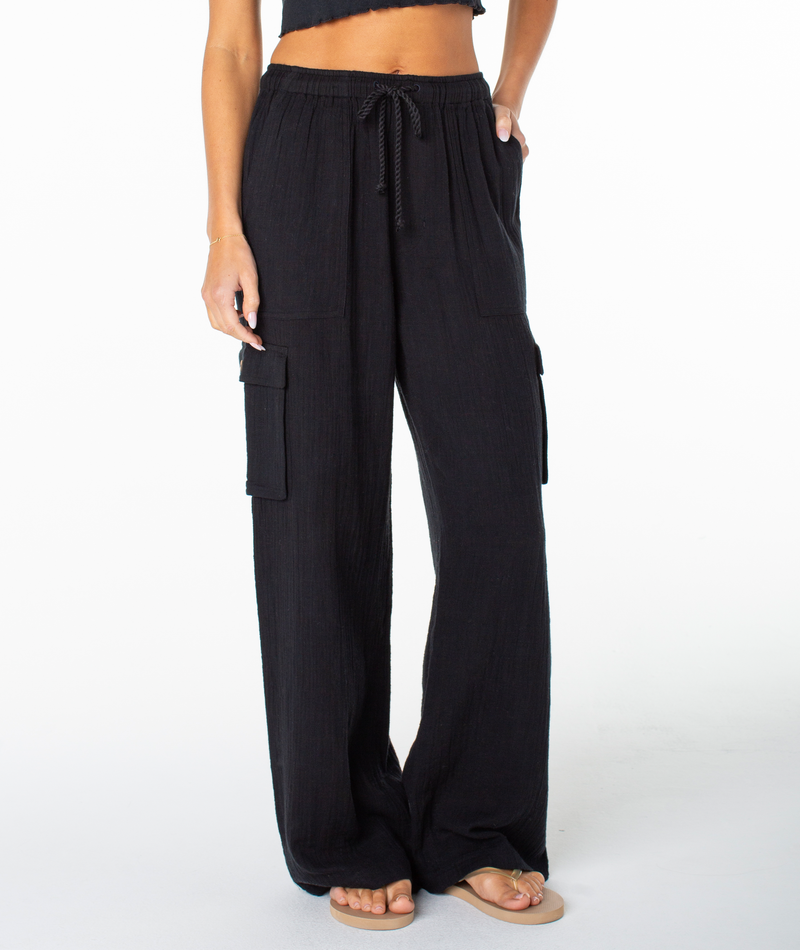 A woman wears the Roxy Precious Cargo Slub Pant in Anthracite Solid, featuring a loose fit with a drawstring waist and side pockets. Styled with flat sandals and a cropped top, she rests her hand in one pocket against a plain white background. These breathable pants from Roxy combine comfort and versatility for everyday wear.