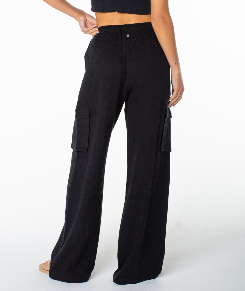 A person stands with one hand on their hip, shown from the back against a white background, wearing the Roxy Precious Cargo Slub Pant in black—these loose cargo pants feature side pockets and are designed for comfort with a breathable fit perfect for everyday wear. The outfit is completed with a black crop top.