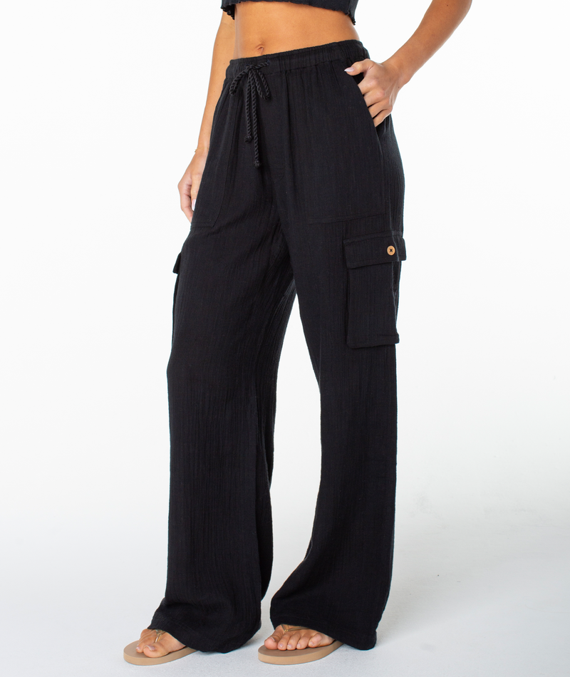 Model showcases the Roxy Precious Cargo Slub Pant—loose black cargo pants featuring a drawstring waist and cargo pockets—paired with a cropped black top and beige sandals. Image is cropped at the shoulders to highlight the outfit against a plain white background.
