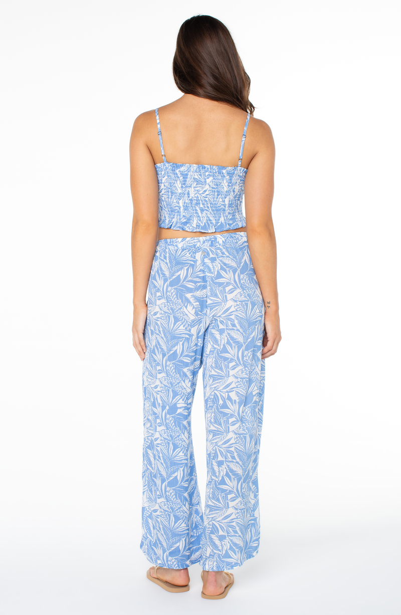 A woman with long brown hair stands barefoot, facing away, wearing the Roxy Hello Sunshine Top in light blue with a white leafy pattern and matching wide-leg pants. The background is plain white.