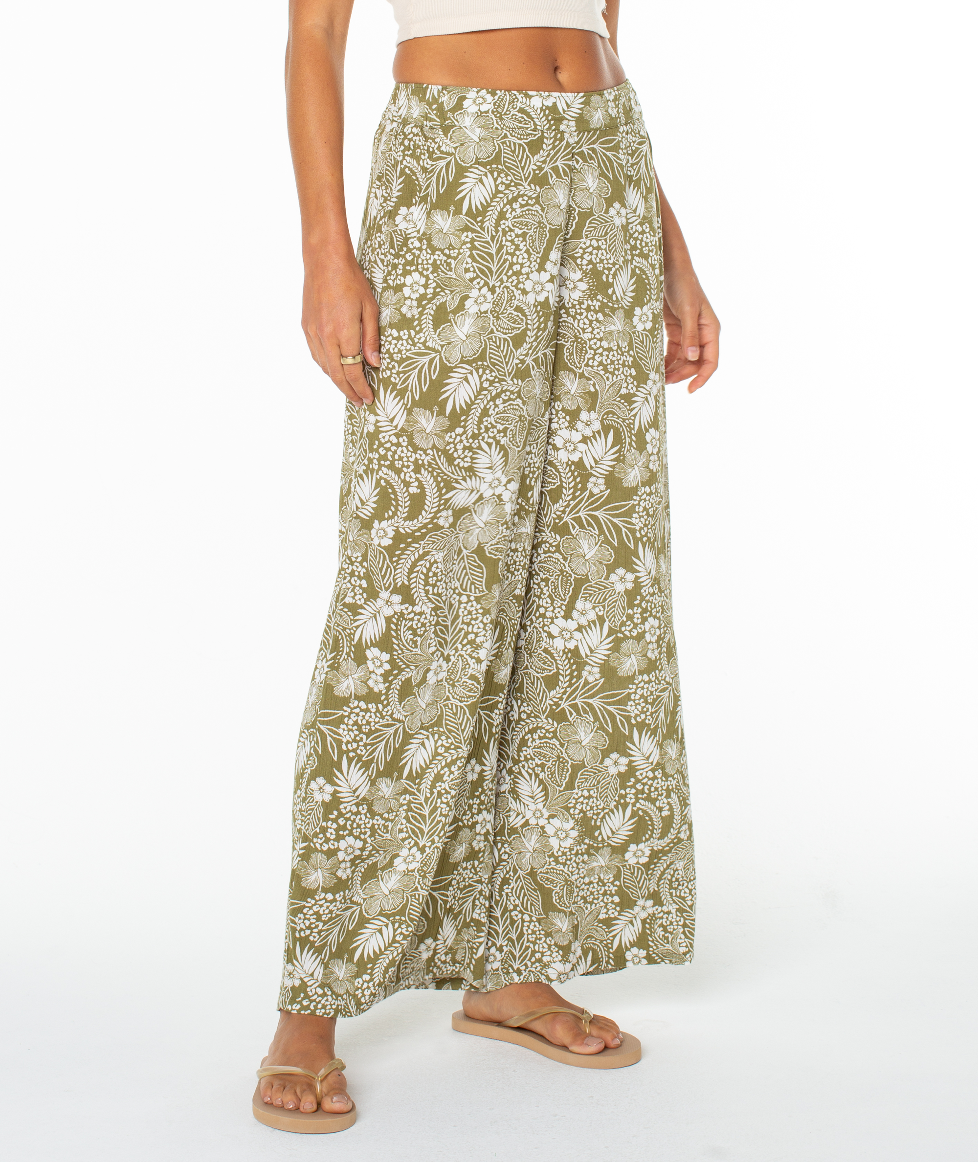 A person wears Roxy's Midnight Ave Pant—olive green, high-waisted with a white floral pattern—paired with a beige crop top and tan sandals, standing against a plain white background.