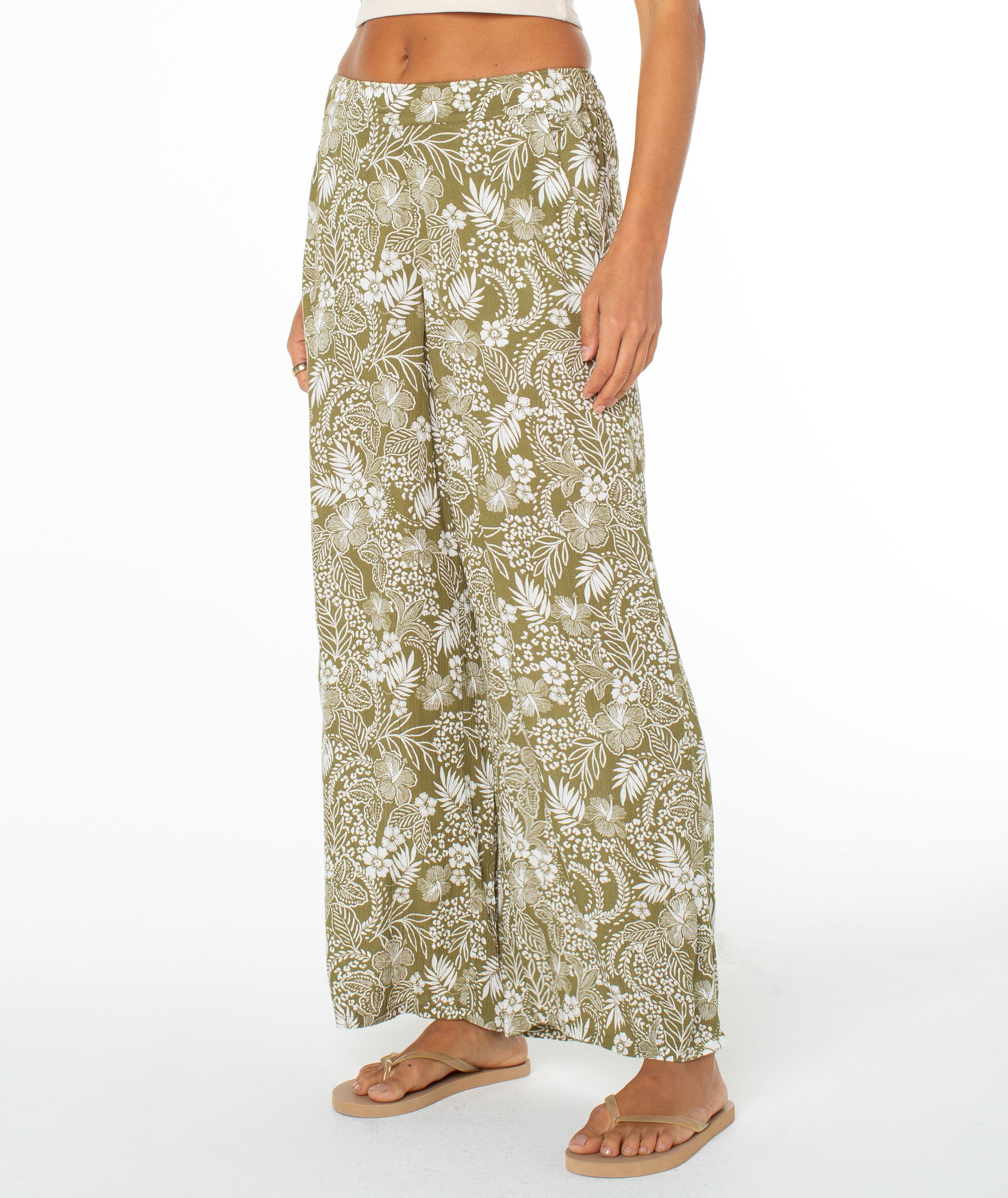 A person models the Roxy Midnight Ave Pant—high-waisted olive green lounge pants with a white tropical leaf print—standing barefoot in flip-flops against a white background.