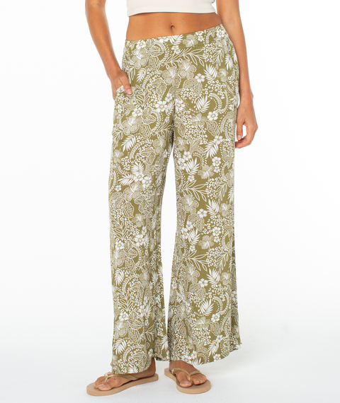 A person wears Roxy’s Midnight Ave Pant—high-waisted, olive green with a white floral and leaf print—paired with a beige crop top and tan sandals, standing against a plain white background.