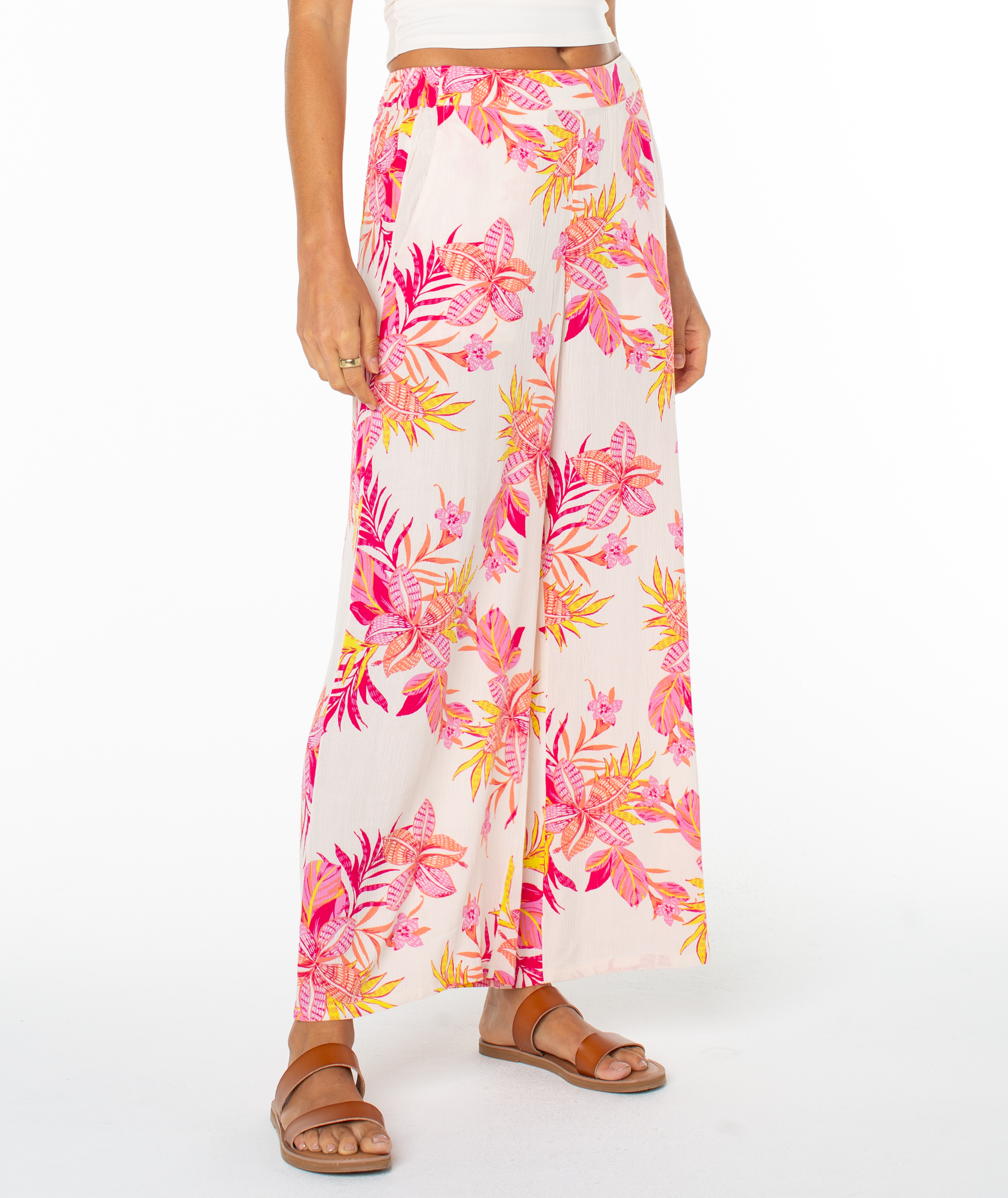 A person in Roxy’s Midnight Ave Pant—wide-leg, high-waisted, with a bold pink, orange, and yellow tropical floral print—pairs it with brown sandals and a white top against a white background, ideal for coastal evenings.