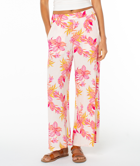 Wearing the Roxy Midnight Ave Pant, featuring a vibrant pink, orange, and yellow floral print, paired with brown sandals—ideal for a breezy coastal night.