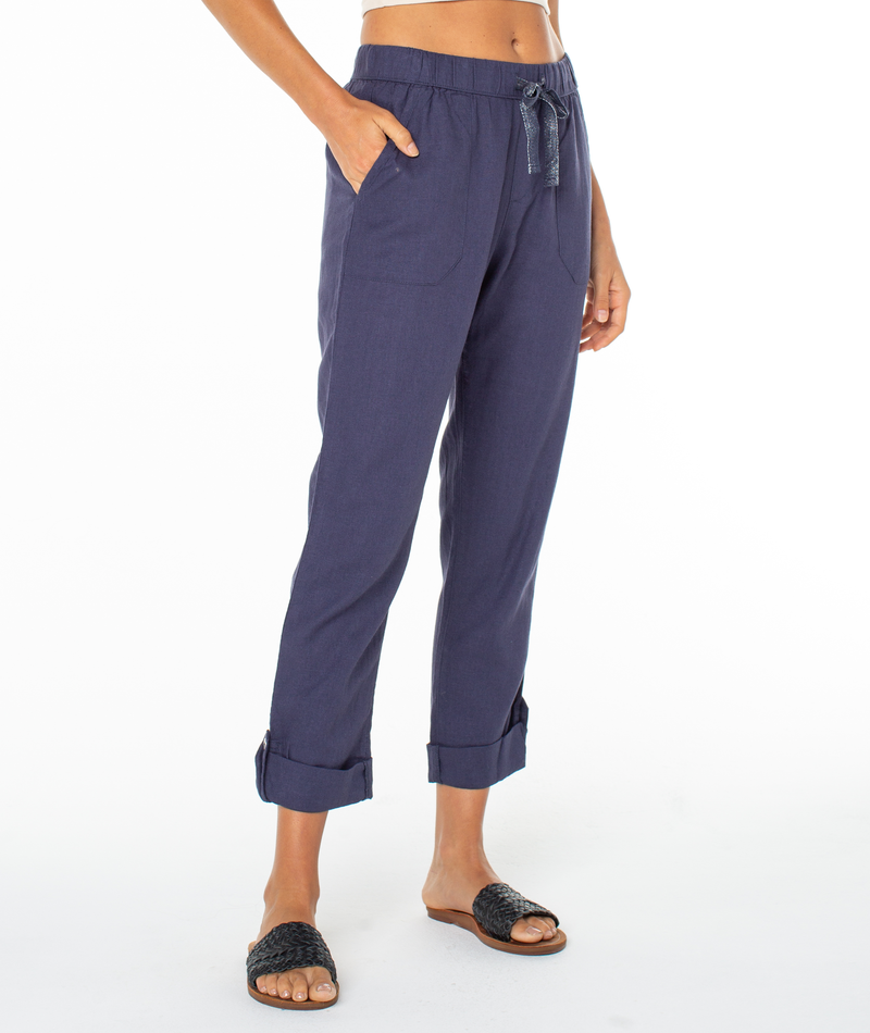 A person wears Roxy's On The Seashore Pant in navy blue linen-viscose with a drawstring waist and hands in pockets, paired with black woven slide sandals, standing against a white background.