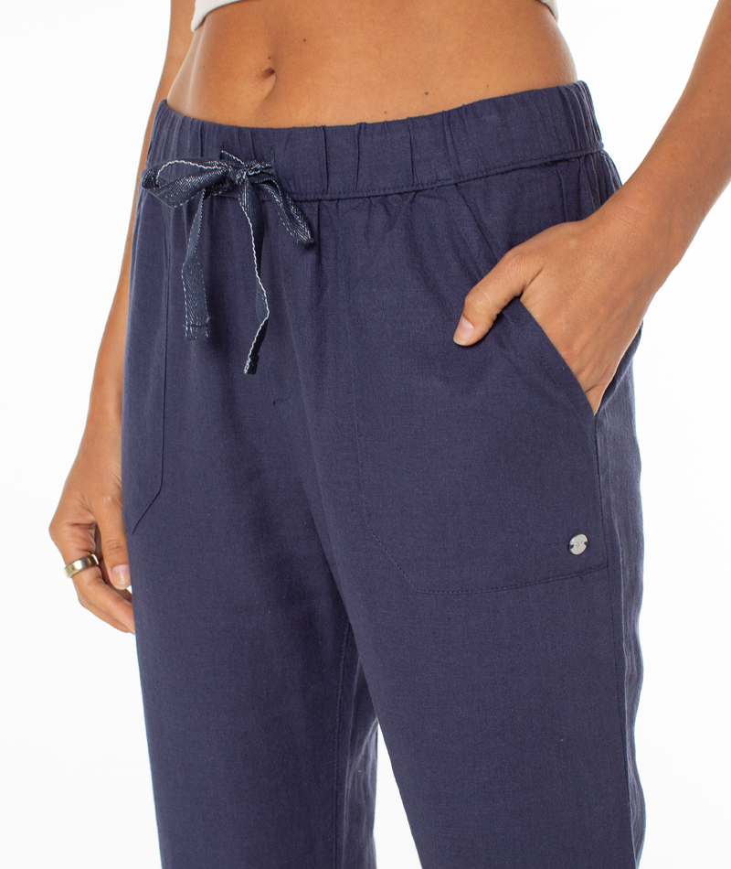 A person wears Roxy's On The Seashore Pant in navy blue linen-viscose, featuring a relaxed straight-leg fit, hands in pockets with a small metallic logo, and a ring on one finger.