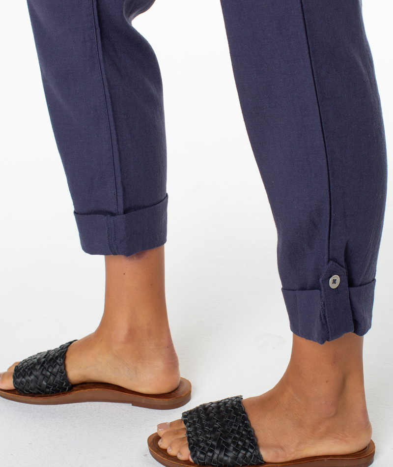 A person wears the Roxy On The Seashore Pant—breathable navy cuffed pants with an elastic waistband—and black woven slide sandals, standing on a white surface. The focus is on their lower legs and feet.