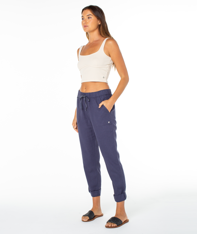 A woman with long brown hair stands against a white background wearing Roxy’s On The Seashore Pant—navy jogger pants with an elastic waistband and rolled cuffs—paired with a cream crop top and black woven slide sandals, hands in her pockets.