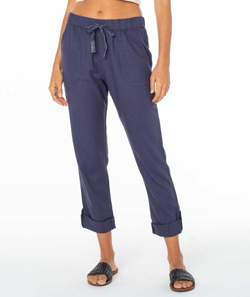 A person wearing Roxy On The Seashore Pant in navy blue with a straight-leg fit, pull-on style, front pockets, rolled cuffs, and black woven slide sandals. The upper body is partially out of frame against a white background.