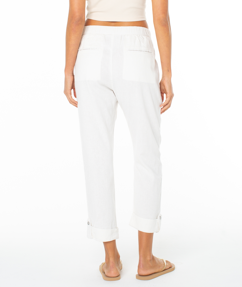 A woman faces away, wearing Roxy's On The Seashore Pant in white with back pockets, a beige crop top, and tan slide sandals against a plain white background.