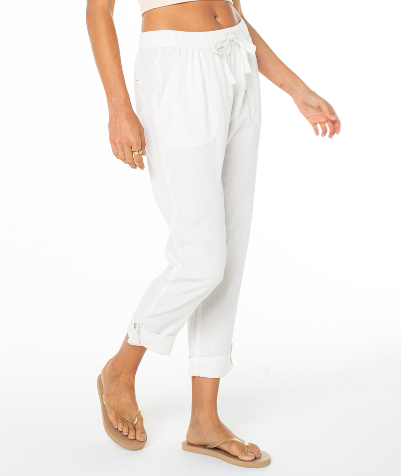 A person wearing Roxy's On The Seashore Pant—white, cuffed, pull-on linen pants with front pockets and a tied waistband—and tan flip-flops stands against a white background.
