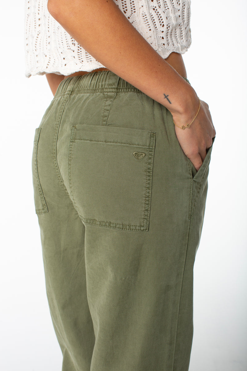 A person wears Roxy’s Sunset Dream Twill Pant in olive green with back pockets and a white knit top, hand in pocket, showing a small cross tattoo and gold bracelet on their wrist.