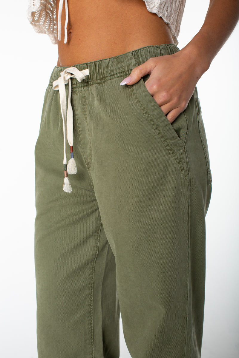 Wearing Roxy Sunset Dream Twill Pant in olive green and a white crochet top, a person stands with one hand in their pocket against a white background.