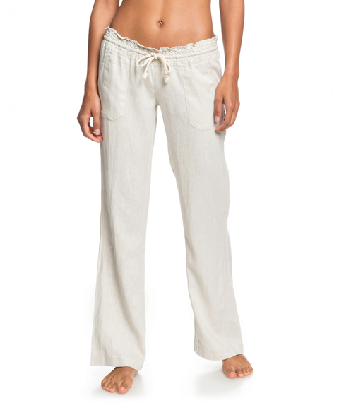 A person stands barefoot against a white background wearing the Roxy Oceanside Pant, a pair of light beige, loose-fitting viscose linen pants with front pockets, a slightly ruffled waistband, and a relaxed flared leg for effortless style.