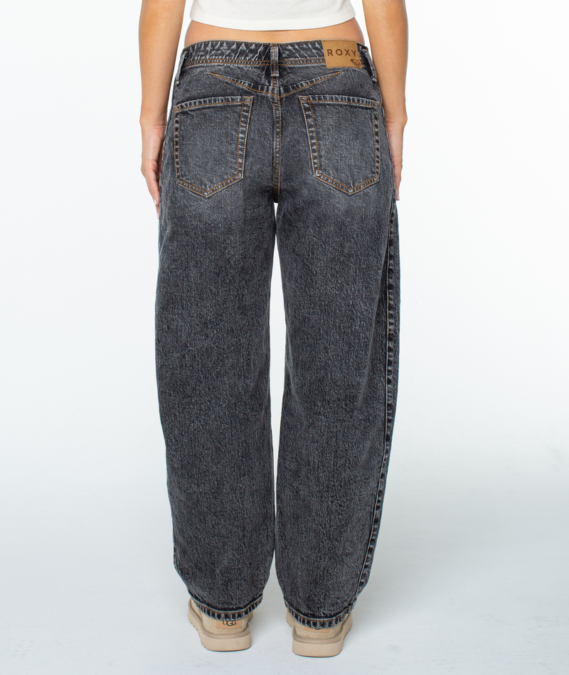 Person stands with their back to the camera, wearing Roxy Surf Pipe Pant—high-waisted, loose-fit black acid wash barrel leg jeans—and beige shoes. The waistband label reads Roxy. Plain white background.