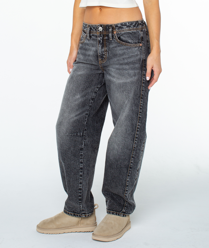 A person models the Roxy Surf Pipe Pant—mid rise, faded black with barrel legs—paired with beige slip-on boots. Only the lower torso and legs are visible against a white background.
