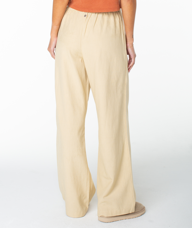 Someone stands on a white background wearing Roxy’s Mavericks Trouser in loose, light beige, a fall must-have, paired with a rust sleeveless top and light-colored shoes. Their left arm is relaxed at their side.