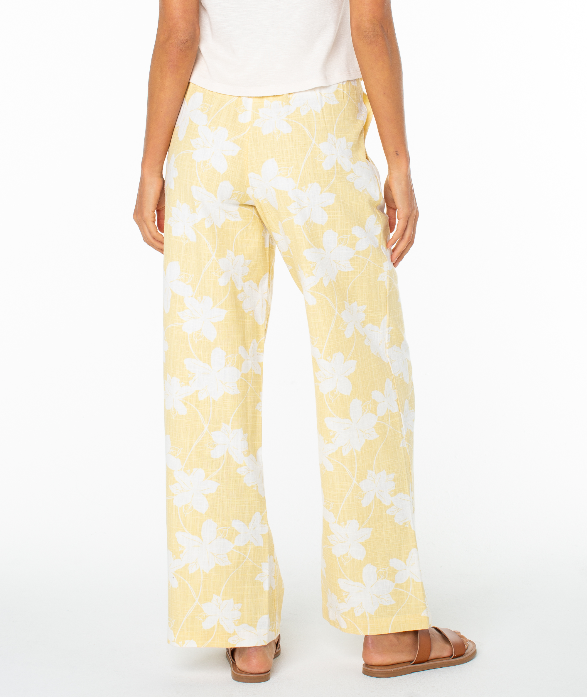 A person faces away against a plain white background, wearing Roxy’s Mavericks Trouser in light yellow with a white floral pattern, paired with a white top and brown sandals.