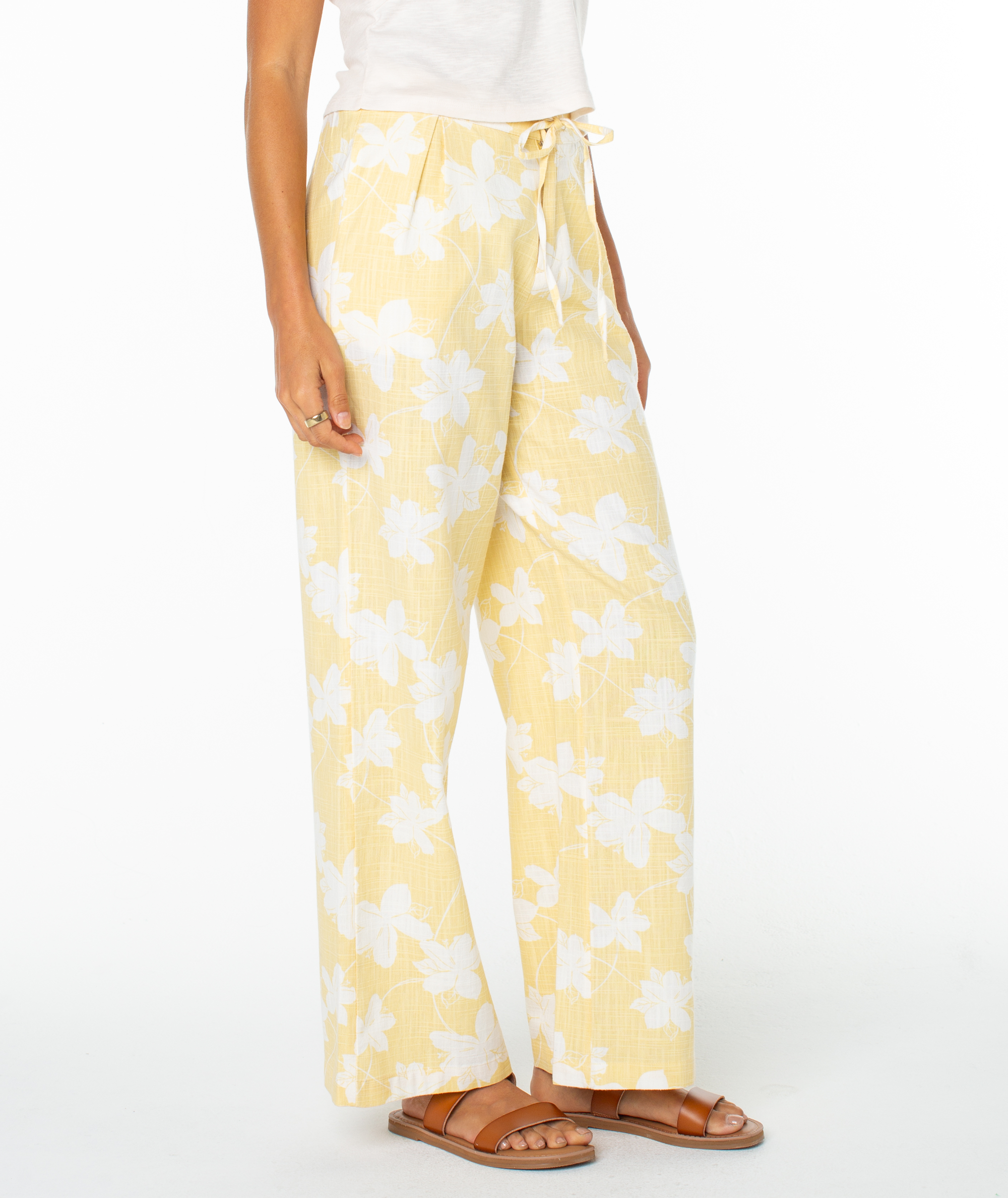 A person stands against a white background in Roxy’s Mavericks Trouser—a light yellow, wide-leg statement pant with a white floral pattern—paired with a white top and brown flat sandals for an effortlessly breezy look.