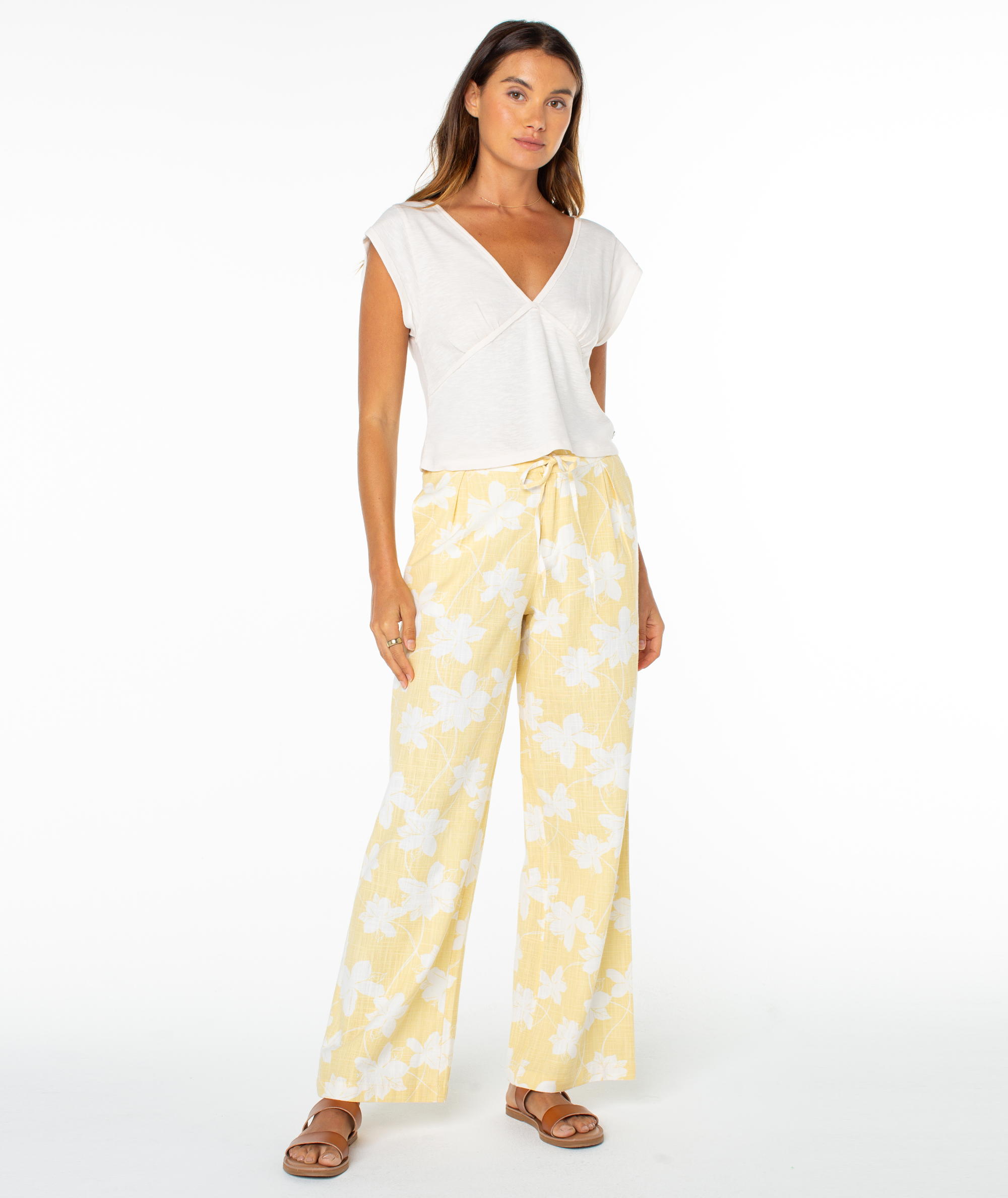 A woman stands against a plain white background wearing a white short-sleeved wrap top, Roxy Mavericks Trouser—a yellow floral statement pant—and brown sandals.