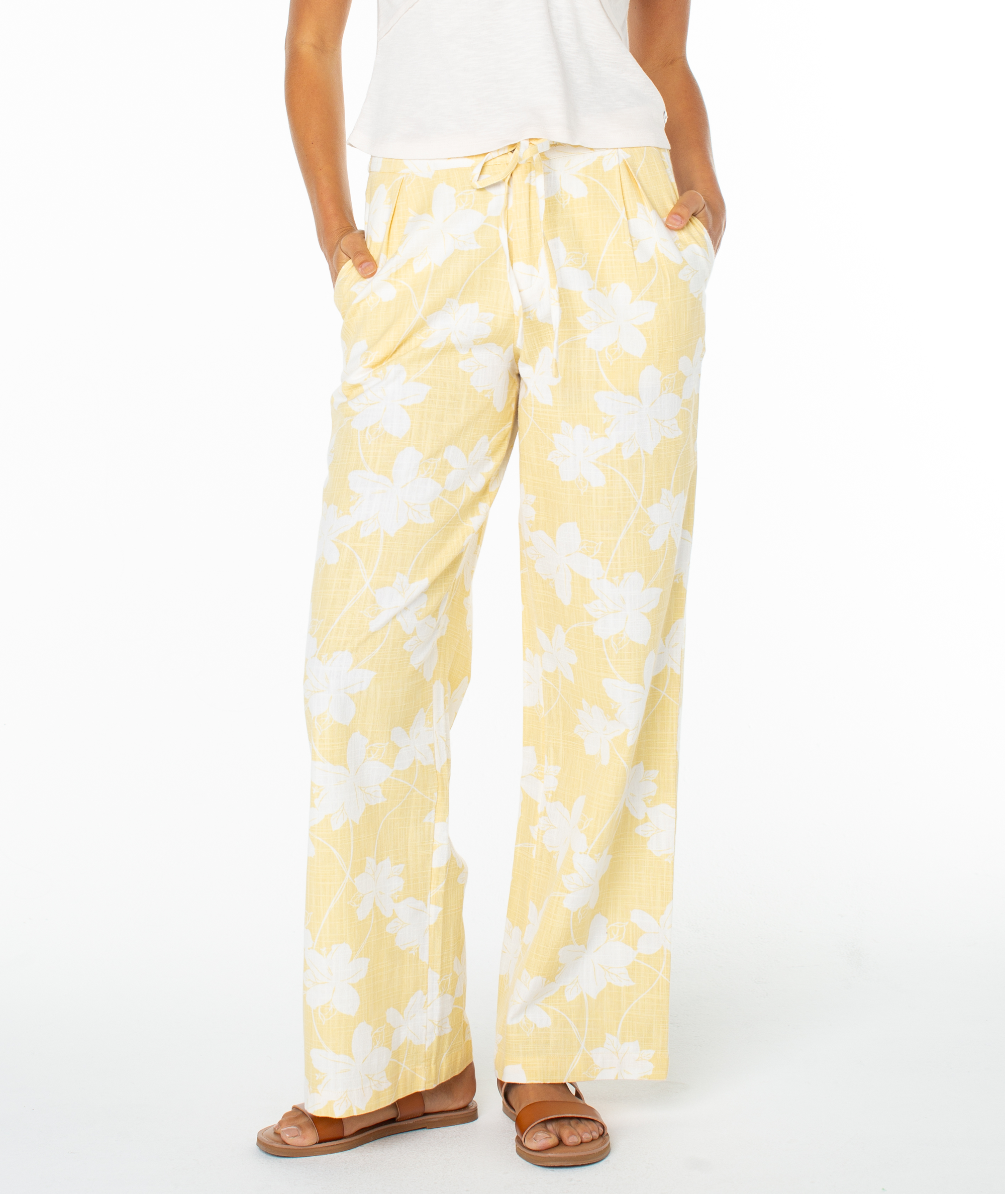 A person wears Roxy’s Mavericks Trouser—light yellow, wide-leg summer pants with a white floral print—hands in pockets, styled with a white top and brown sandals against a plain white background. Ideal for warm-weather looks.