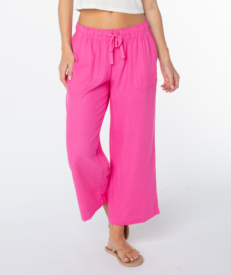 A person in Roxy’s bright pink Crop Lagoon Pant with oversized pockets and a white cropped top, styled with beige sandals, stands against a plain white background.