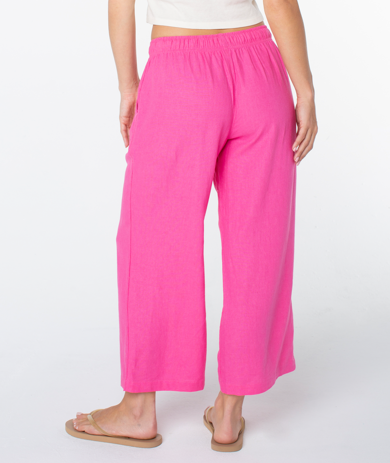 Someone stands facing away from the camera against a plain white background, wearing Roxy's bright pink Crop Lagoon Pant with oversized pockets, paired with a white top and beige flip-flops.
