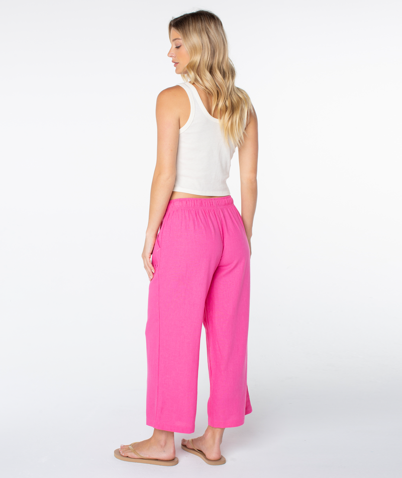 A woman with long blonde hair wears a white sleeveless top, Roxy Crop Lagoon Pant in bright pink with oversized pockets, and beige flip-flops. She stands looking left against a plain white background.