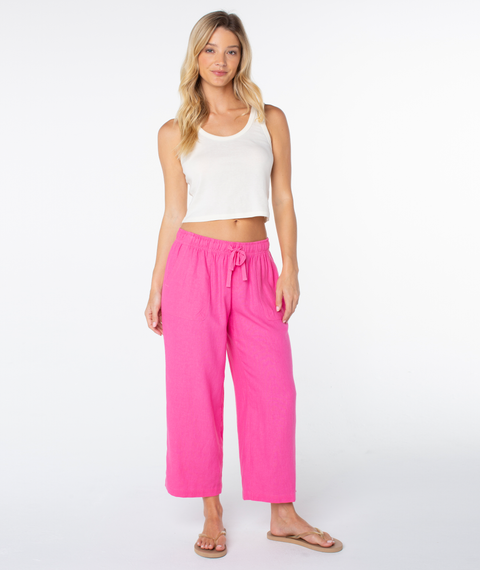 A woman stands against a white background, wearing a white sleeveless crop top and Roxy's bright pink Crop Lagoon Pant in a linen blend with oversized pockets, paired with beige sandals. Her long blonde hair completes the casual look.