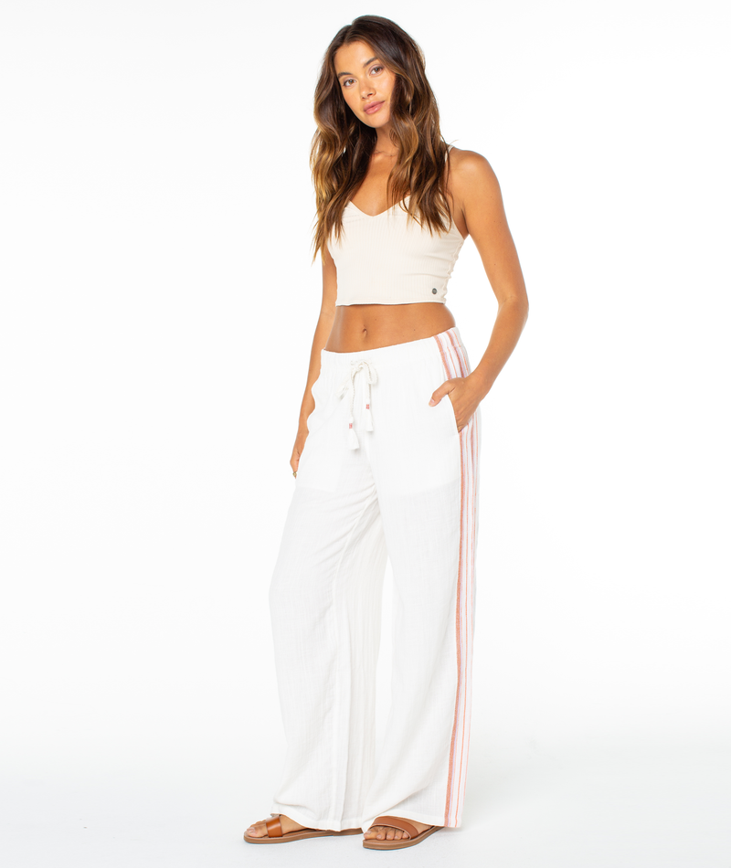 A woman with long, wavy brown hair poses casually against a white background wearing a beige crop top, Roxy Fiesta Siesta Pant with pink side stripes, and brown sandals.