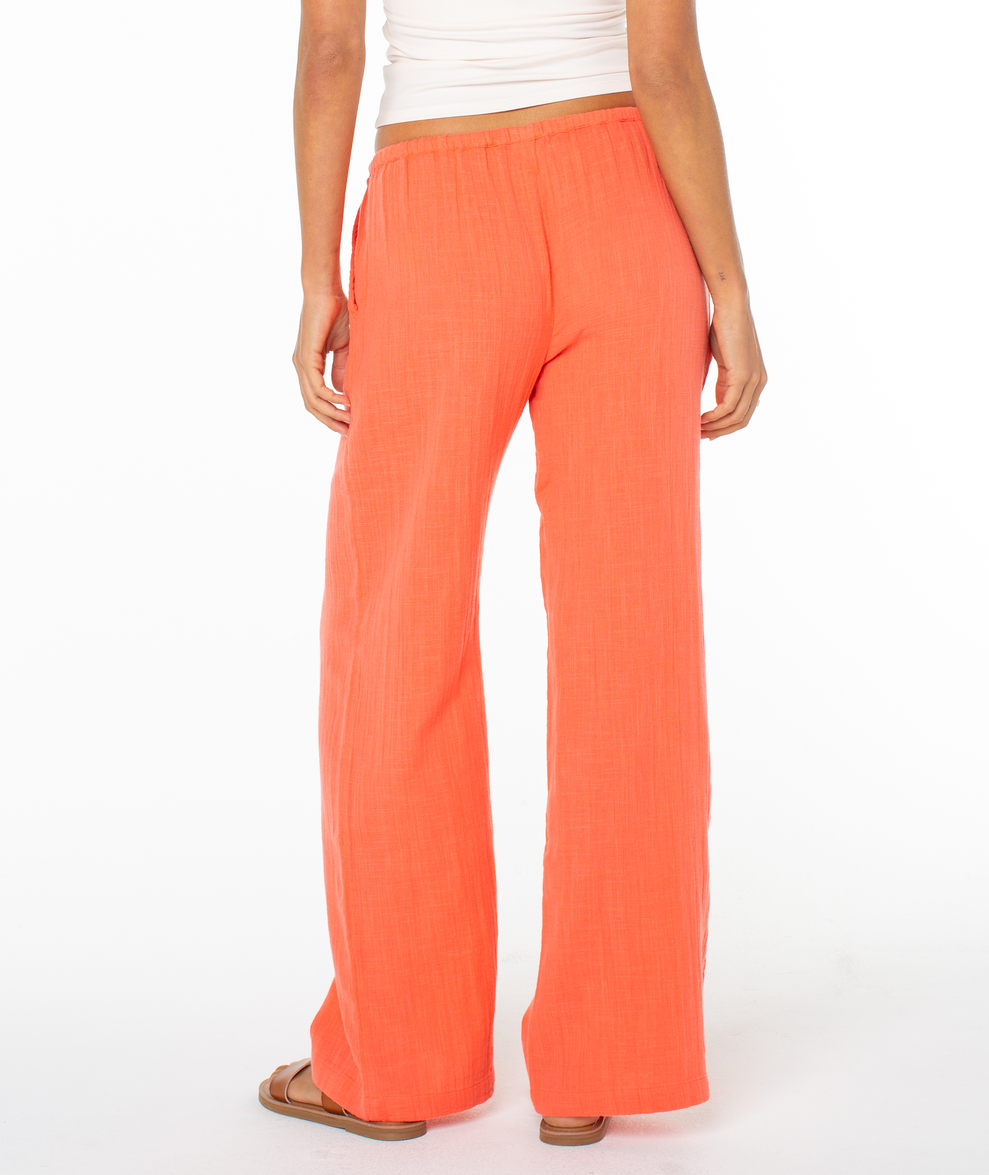 A person seen from the back wears loose-fitting, bright orange Roxy Iyla Pant 2 with an elastic waistband, paired with a white sleeveless top and brown sandals. The relaxed fit pants stand out against the plain white background.