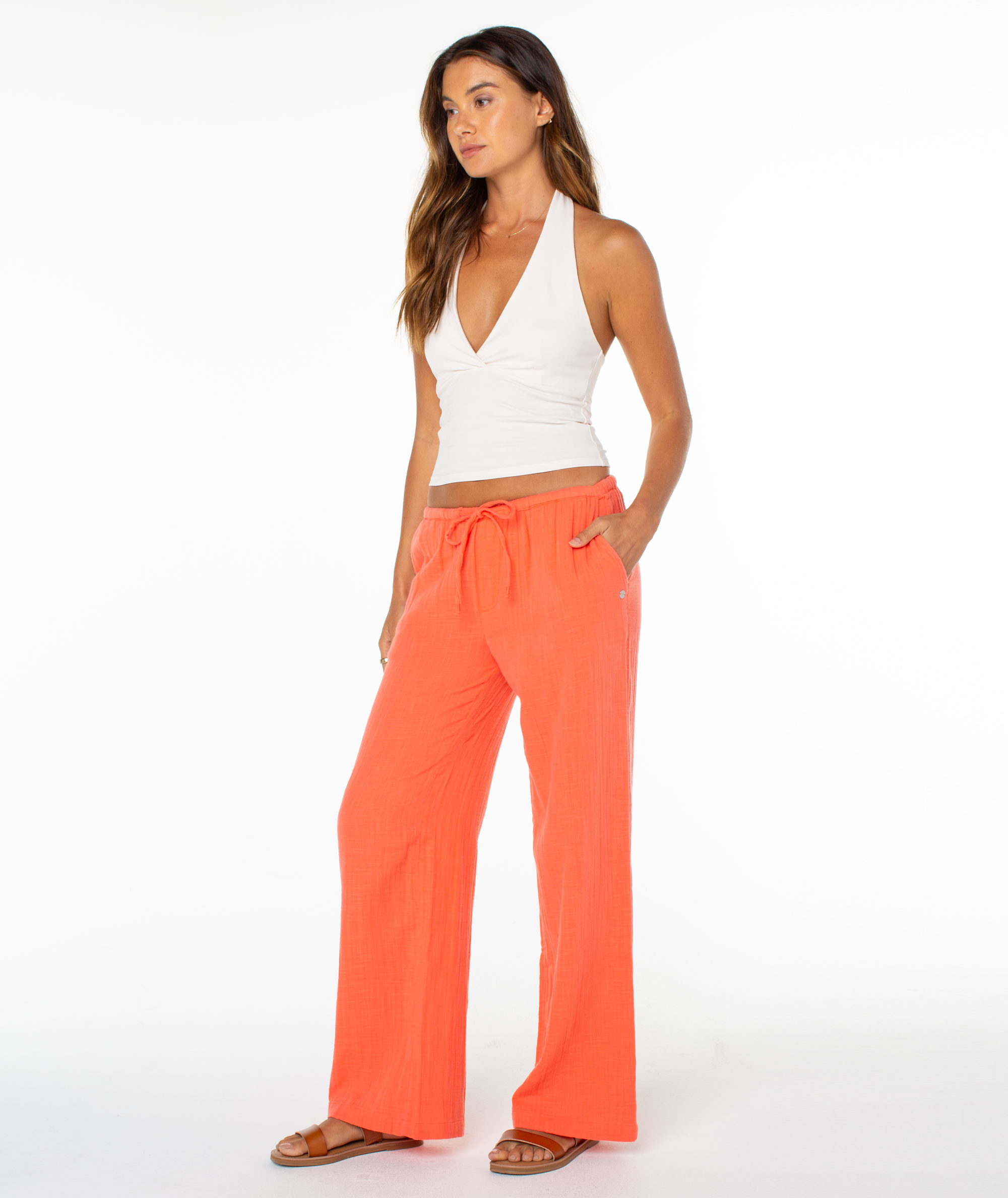 A woman stands against a white background wearing a white halter top, Roxy's relaxed fit Iyla Pant 2 in bright orange with a drawstring waist, and brown sandals—embodying effortless coastal style with her long hair and hands in her pockets.