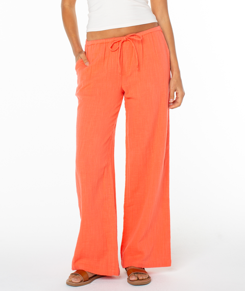A person wears Roxy’s bright orange Iyla Pant 2 with a relaxed fit and elastic waistband, paired with a white sleeveless top and brown sandals, standing against a white background for an effortless coastal look.