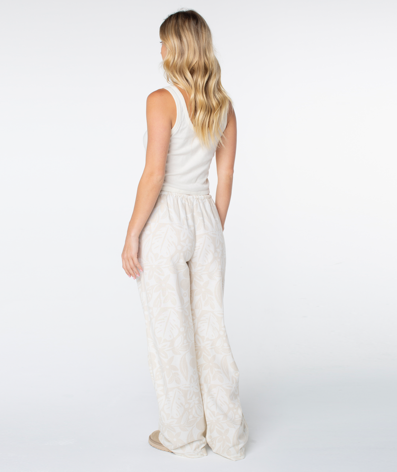 A woman viewed from behind, wearing a cream-colored tank top and Roxy Iyla Pant 2 in a light tone with a subtle leaf pattern.